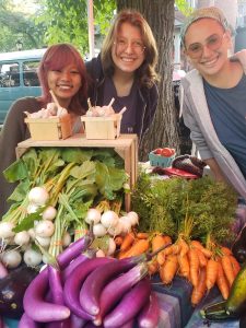 Three smiling people behind display of turnips, carrots, and eggplants